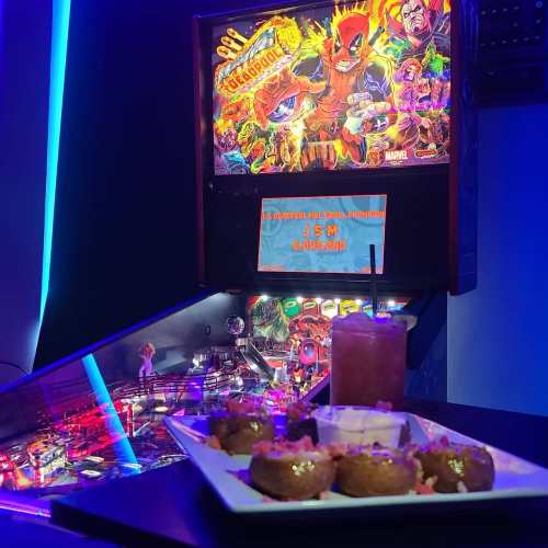 A pinball machine featuring Deadpool, with a plate of food and a drink in the foreground, set in a vibrant arcade.