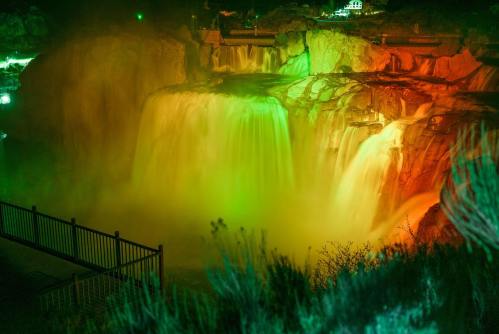 A vibrant waterfall illuminated in green and orange lights, surrounded by mist and a railing in the foreground.