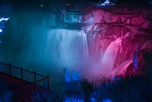 A vibrant waterfall illuminated in pink and blue lights, surrounded by mist and a dark landscape.