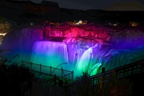 A vibrant waterfall illuminated with colorful lights at night, surrounded by a scenic landscape and viewing platform.