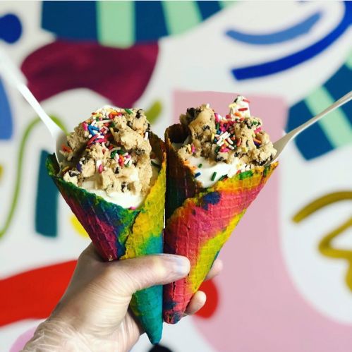 Two colorful ice cream cones topped with cookie dough and sprinkles, held against a vibrant, abstract background.
