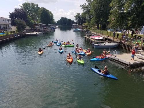 A group of people kayaking on a calm river, surrounded by trees and houses, with others enjoying the waterfront.