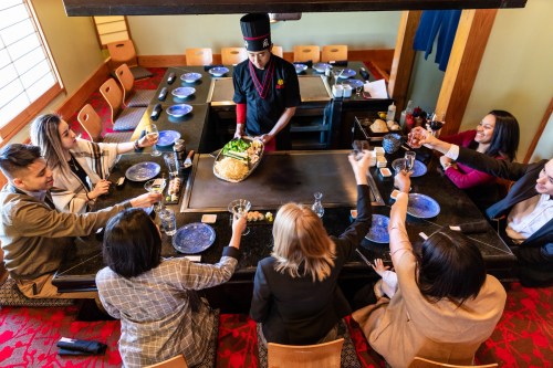 A chef serves food at a teppanyaki table while guests raise glasses in celebration.