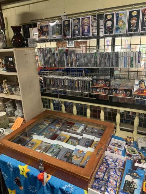 A display case filled with collectibles, surrounded by shelves of DVDs and figurines in a shop setting.