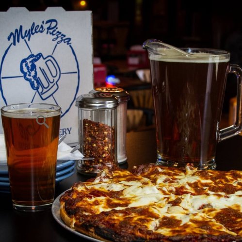 A delicious pizza on a plate, accompanied by a pitcher of beer, a glass of beer, and a shaker of red pepper flakes.