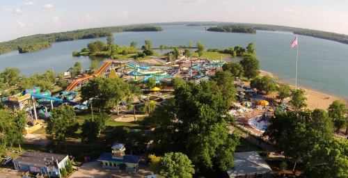 Aerial view of a water park by a lake, featuring slides, pools, and beach areas surrounded by greenery.