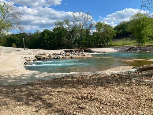 A serene river scene with clear water, rocky banks, and lush green trees under a blue sky with fluffy clouds.