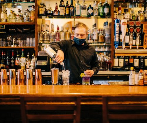 A bartender in a black shirt and mask pours a drink at a well-stocked bar, surrounded by various bottles.