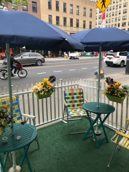 A cozy outdoor seating area with colorful umbrellas, chairs, and flower baskets, overlooking a city street.