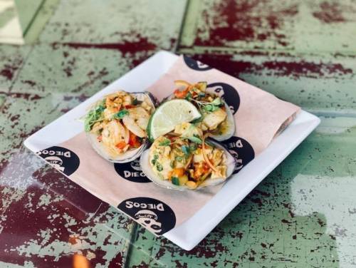 Three tacos topped with colorful vegetables and lime, served on a plate with a branded paper liner.