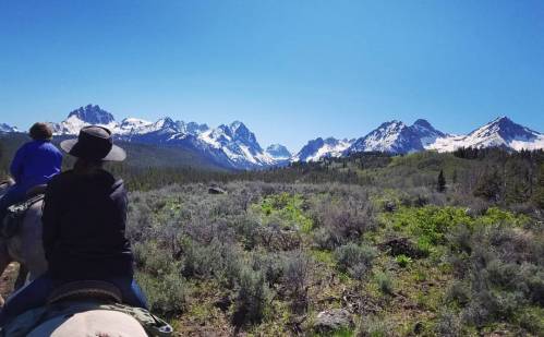 Two riders on horseback overlook a scenic mountain range with snow-capped peaks under a clear blue sky.