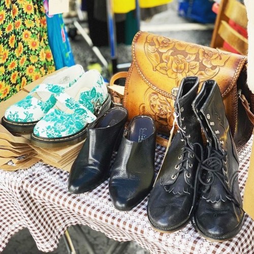A display of various shoes, including floral clogs, black ankle boots, and a leather handbag on a checkered tablecloth.