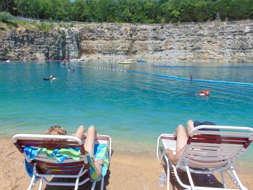 Two people relax on beach chairs by a clear blue lake, with swimmers and a rocky cliff in the background.