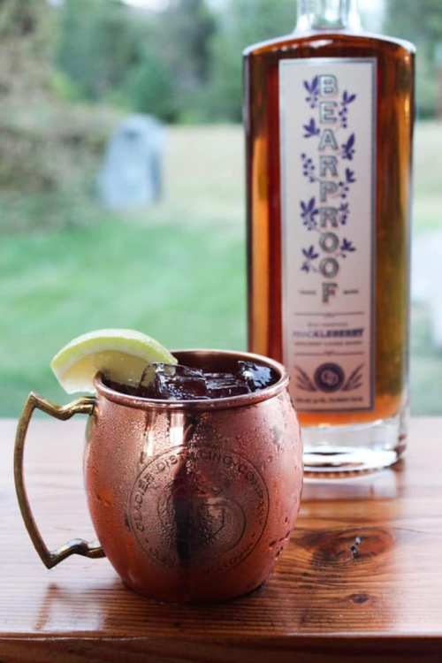 A copper mug filled with a dark drink and a lime wedge, next to a bottle of Bearproof blueberry liquor.