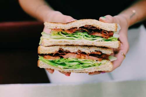 A person holds a stacked sandwich with layers of lettuce, tomato, and bacon between slices of bread.