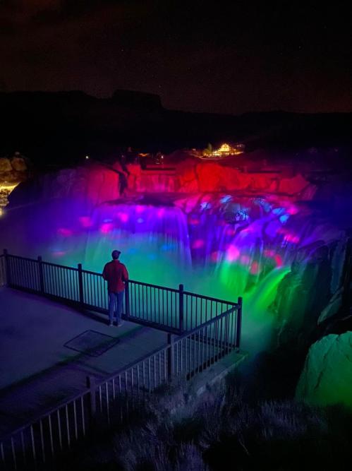 A person stands by a railing, gazing at a brightly lit waterfall with colorful lights at night.