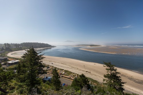 A scenic view of a river meeting the ocean, surrounded by trees and sandy beaches under a clear blue sky.