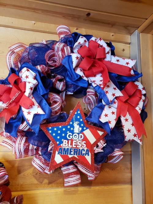 A patriotic wreath featuring red, white, and blue ribbons, bows, and a star with "God Bless America" in the center.