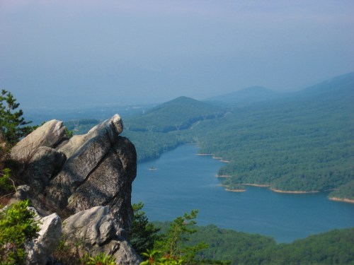 A rocky outcrop overlooks a serene lake surrounded by lush green mountains under a clear blue sky.