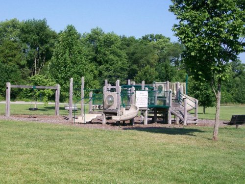 A playground with slides, swings, and climbing structures, surrounded by grass and trees under a clear blue sky.