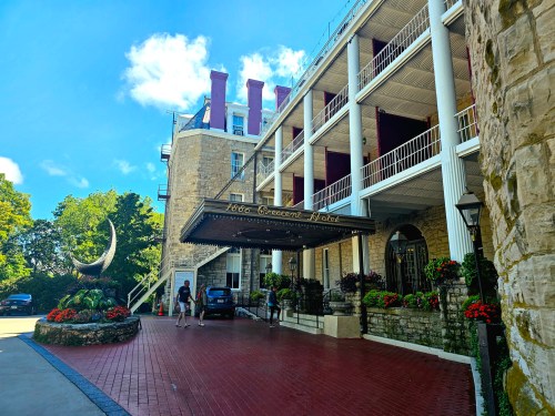 Historic hotel entrance with stone architecture, red brick driveway, and colorful flower beds under a blue sky.