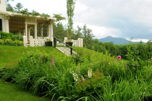 A lush garden with colorful flowers, leading to a white gazebo and a backdrop of green mountains under a cloudy sky.