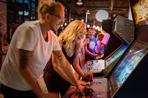 Two people joyfully playing vintage arcade games in a lively, retro-themed arcade filled with others.