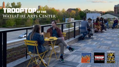 A rooftop scene at the WaterFire Arts Center with people enjoying drinks and snacks at yellow tables.