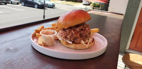A pulled pork sandwich on a bun with coleslaw, served with fries and a side of dipping sauce on a white plate.