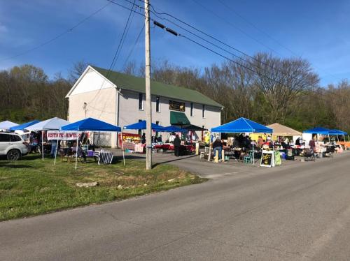 A lively outdoor market with colorful tents set up along a road, next to a large building and surrounded by greenery.