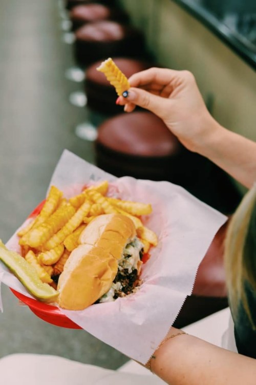 A hand holding a crinkle-cut fry above a plate with two hot dogs, fries, and a pickle.