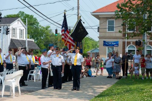 A group of uniformed individuals saluting at a public event, with spectators and American flags in the background.