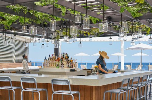 A stylish outdoor bar with hanging lanterns and greenery, featuring a bartender in a hat and a view of the ocean.