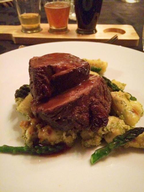 A plate of tender beef filet on a bed of gnocchi and asparagus, drizzled with sauce, with drinks in the background.