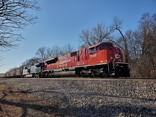 A red Canadian Pacific locomotive pulls freight cars along a railway track, surrounded by bare trees and a clear blue sky.