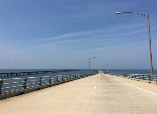 A long, straight bridge extends over calm blue waters under a clear sky, with streetlights lining the sides.