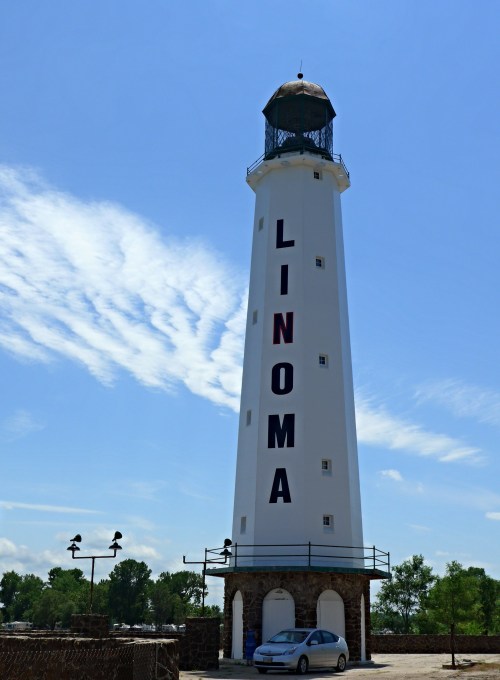 A tall white lighthouse with "LINOMA" written in large letters, set against a blue sky with wispy clouds.