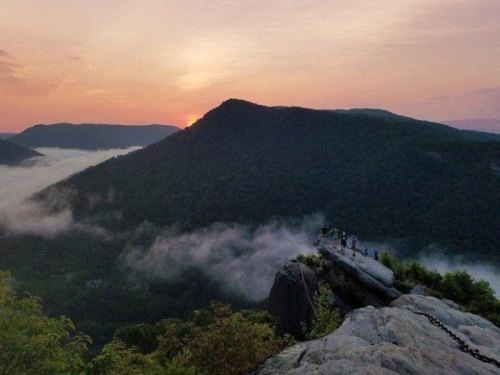 A group of people stands on a rocky outcrop overlooking a misty valley at sunset, with mountains in the background.