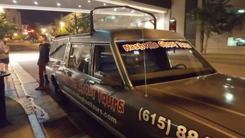 A dark vehicle with "Nashville Ghost Tours" signage parked on a city street at night, with people nearby.
