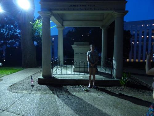 A person stands near a monument at night, illuminated by a light, with a small American flag nearby.