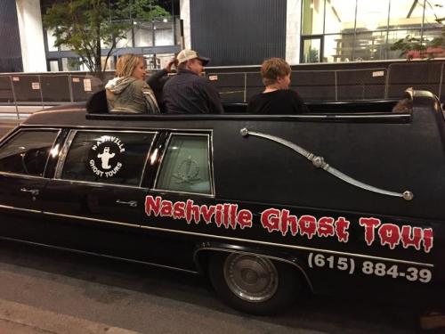 A group of people sitting in a black hearse with "Nashville Ghost Tour" signage parked on a city street at night.