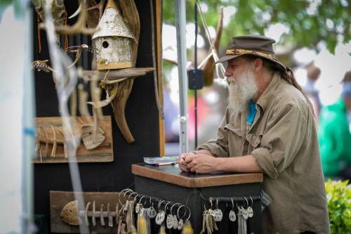 A bearded man in a hat stands at a booth displaying handmade wooden crafts and jewelry at an outdoor market.