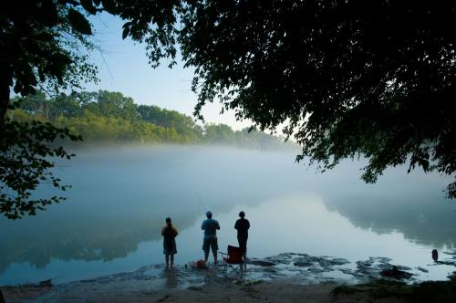 Three people fishing by a misty river at dawn, surrounded by trees and calm water.