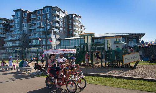 A vibrant park scene with families enjoying a playground and people riding a multi-seater bike near modern buildings.