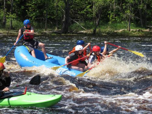 A group of people in a blue raft navigating rapids, with kayakers nearby on a sunny day.