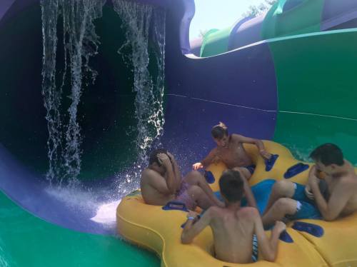 A group of four boys in a yellow inflatable raft, splashing water as they exit a colorful water slide.