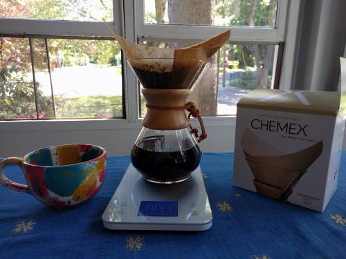 A Chemex coffee maker brews coffee on a scale, with a colorful mug and a box of Chemex filters nearby.