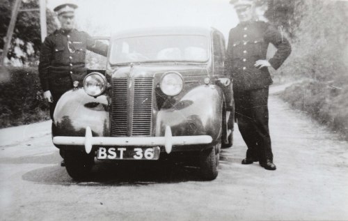 Two police officers in uniform stand beside a vintage car on a rural road, with a license plate reading "BST 36."