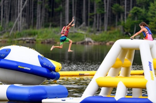 A child jumps off an inflatable water structure into a lake, while another child plays on a nearby inflatable.