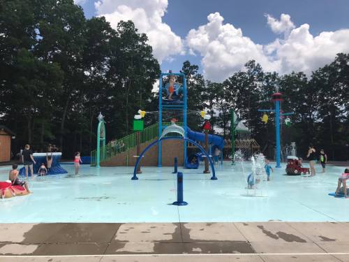 A vibrant water park scene with children playing in a splash area, slides, and water features under a blue sky.